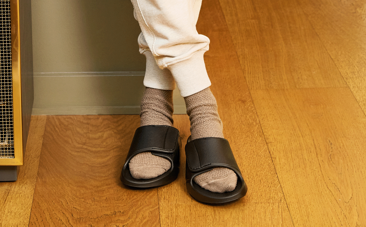 Person wearing black slides with brown socks on a wooden floor.