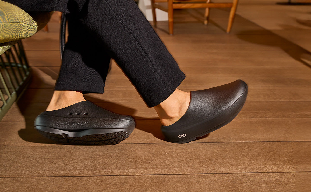 Person wearing black oofos clog plus shoes on a wooden floor