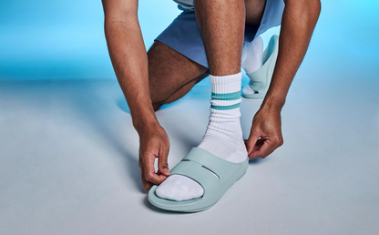 Men's OOahh Slide Sandal - Mist model adjusting shoe in studio with blue background | lifestyle