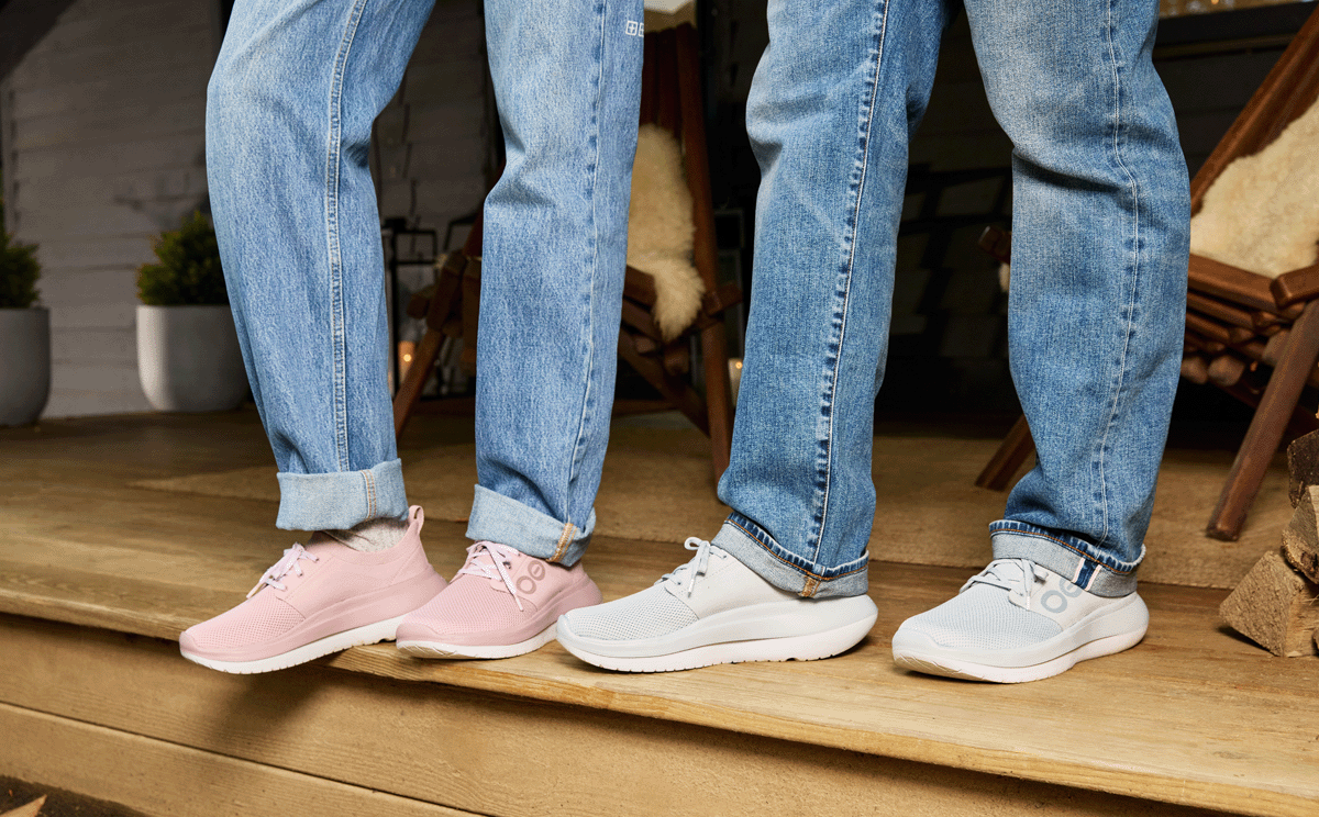 Two pairs of legs wearing blue jeans and different colored sneakers on a wooden floor.