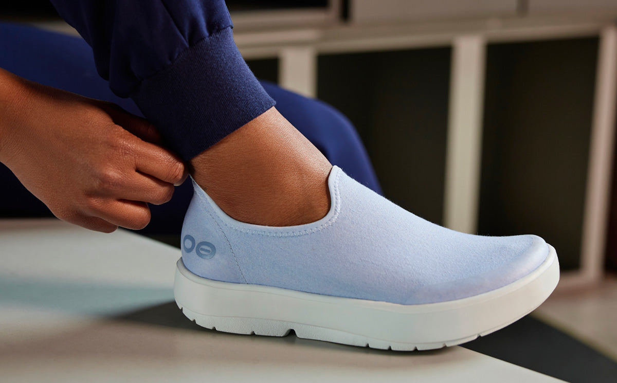 Woman wearing nurse scrubs, pulling on OOmg eeZee Low Shoe in Purple Jade in front of lockers
